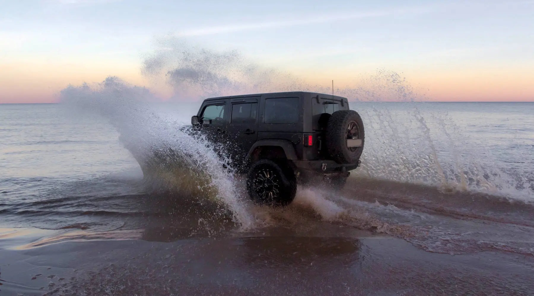 A black Jeep on the beach splashing water as it drives through the shoreline. The sun is setting in the background.