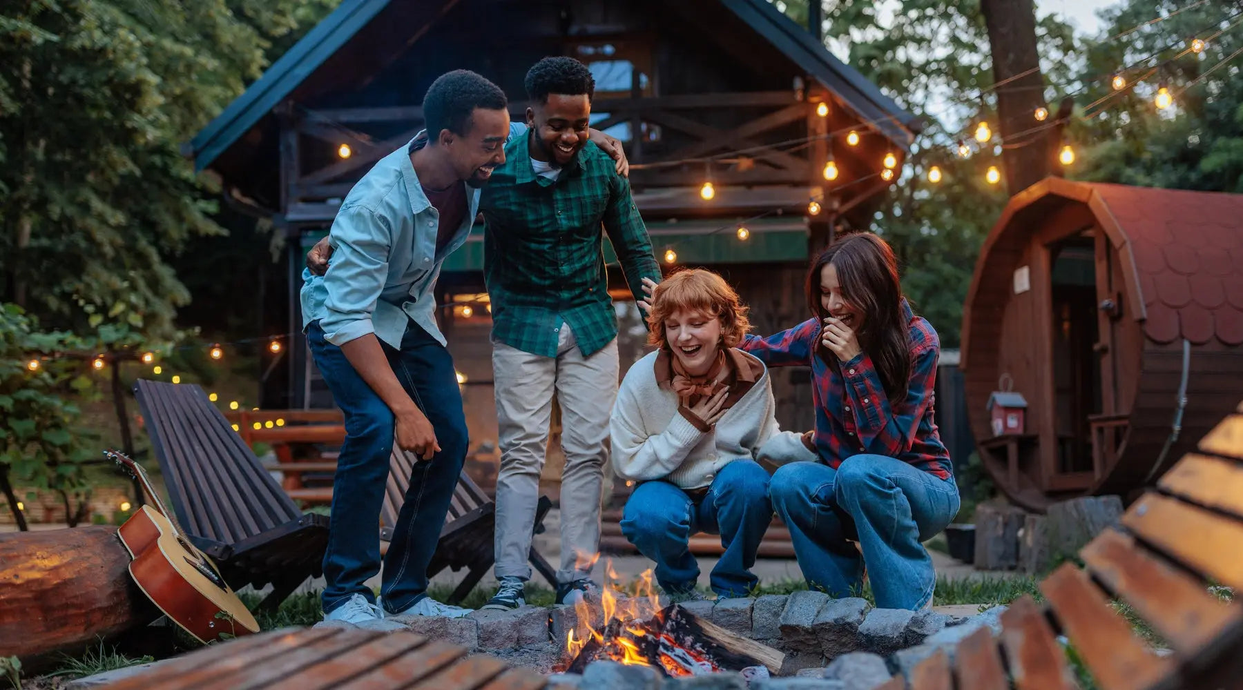 Four people gather around a fire pit. Two are crouching and two are standing. Behind them is a log cabin with fairy lights.