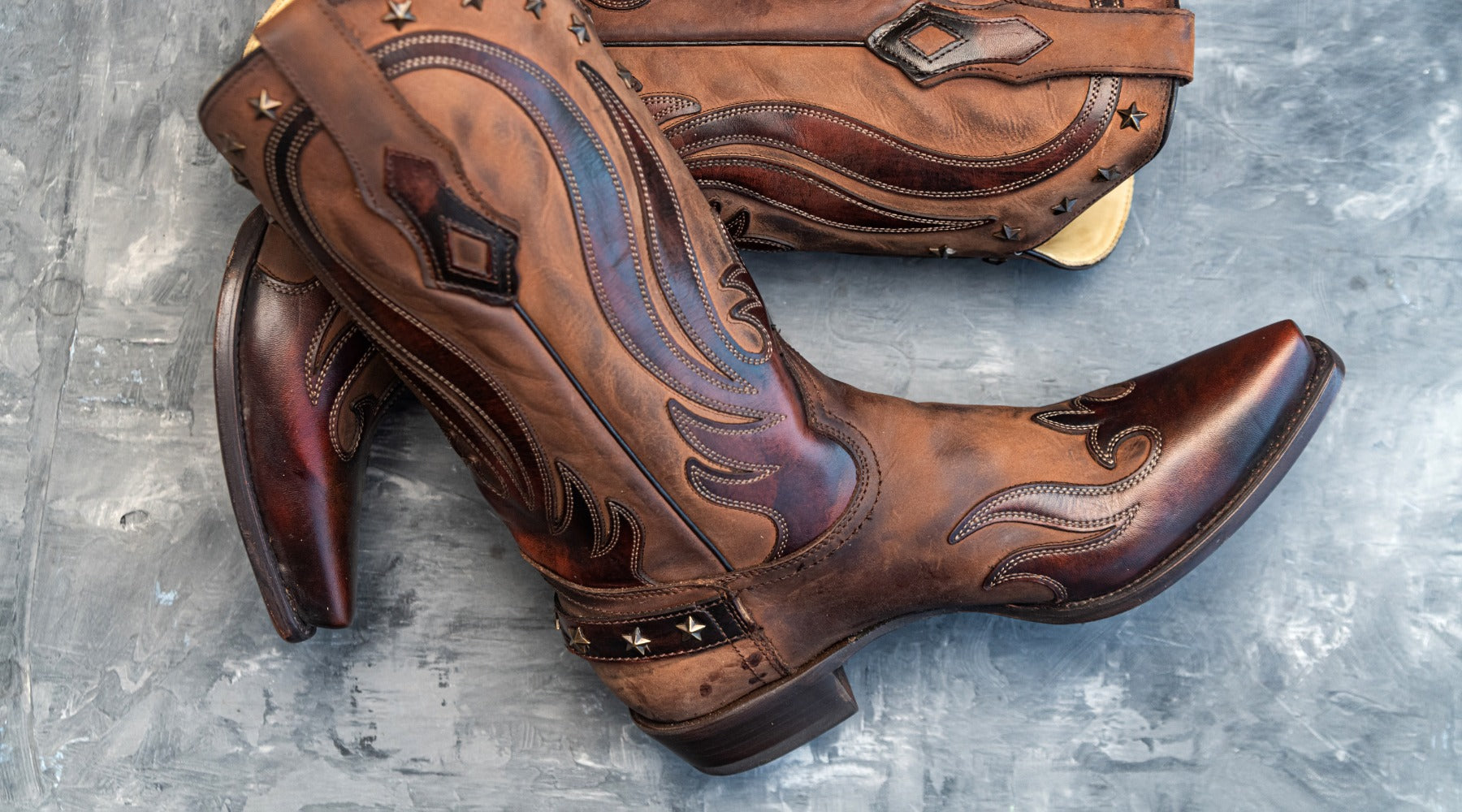 A close-up of a pair of cowboy boots made with brown cowhide leather with decorative trim on a concrete floor.