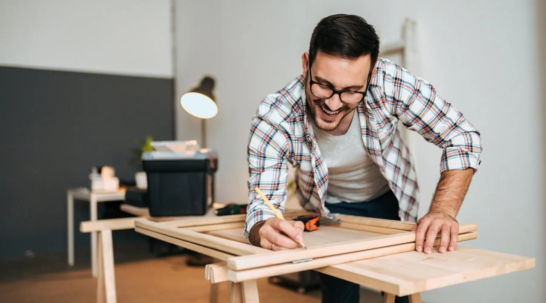 A smiling man leaning over a work surface and using a pencil to make marks on a wooden DIY project.