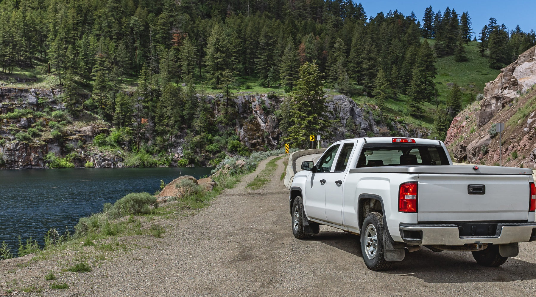 A white pickup truck parked on a dirt road along a mountain, next to a river surrounded by trees on a sunny day.