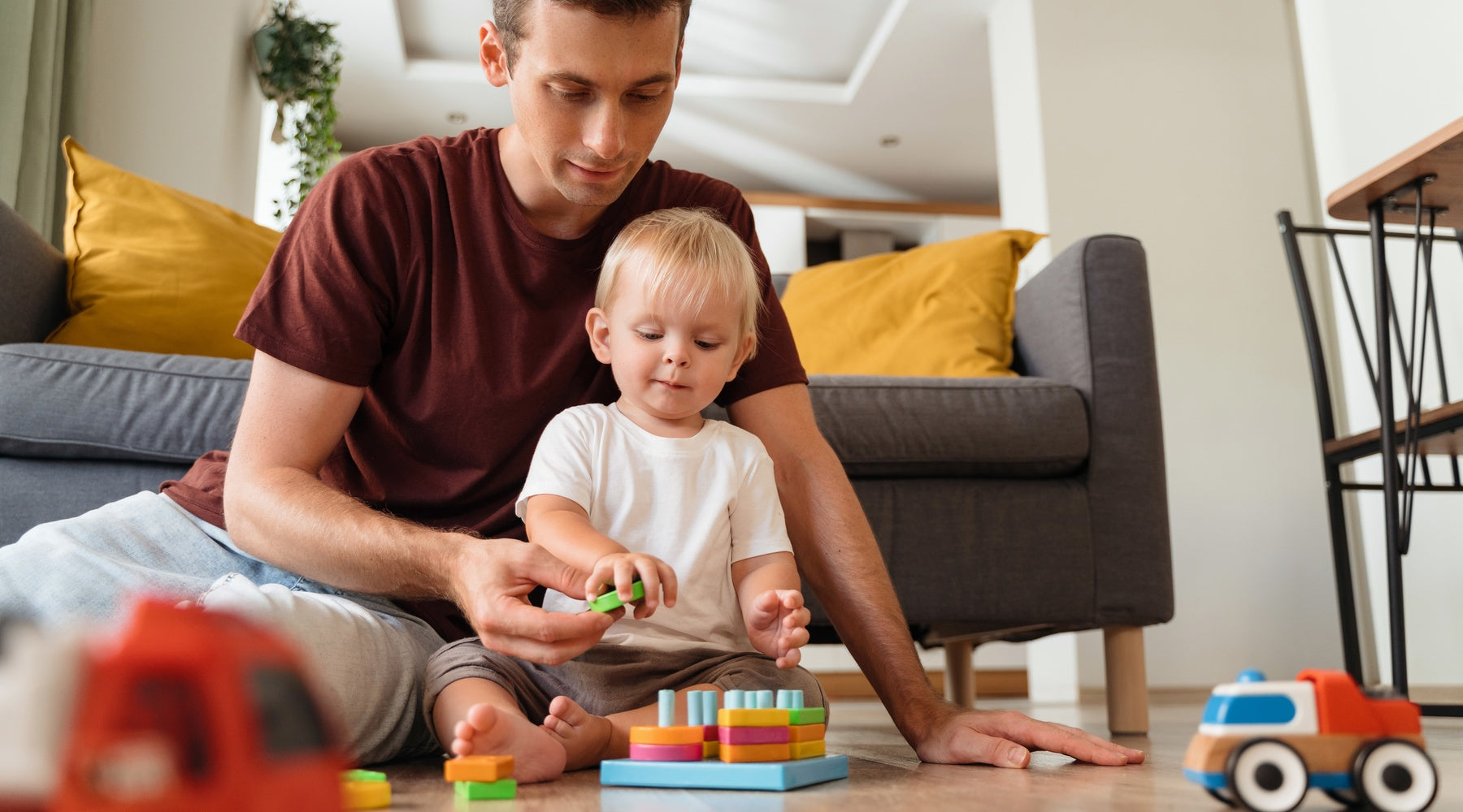 A father sits with his toddler on the living room floor and helps him place blocks on a toy. Other toys surround them.