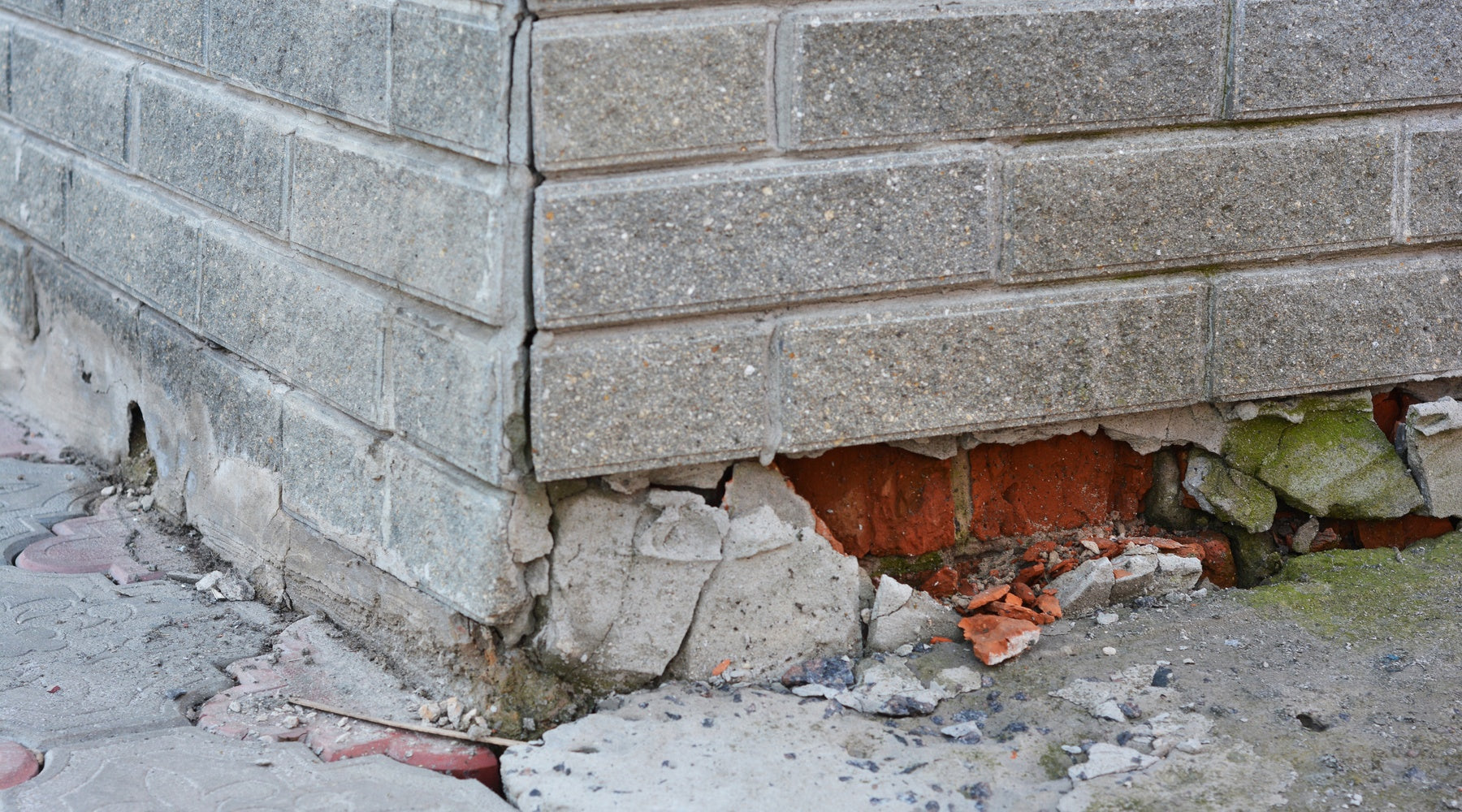 The corner of a house made of gray bricks. The foundation is crumbling away at the bottom of the house.