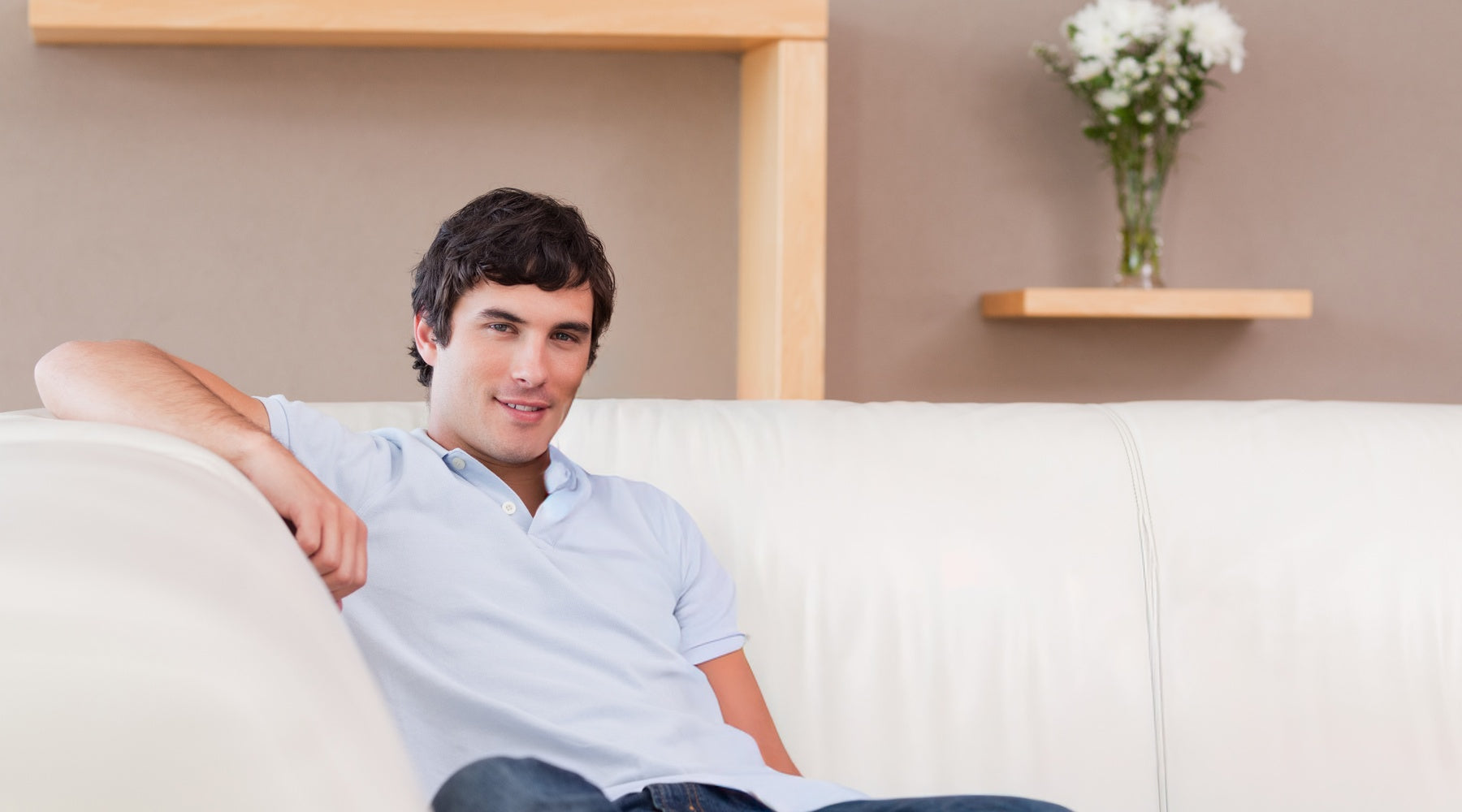 A man reclines on a white sofa in a minimalist living room with wooden shelves, folded cloth, and white flowers.