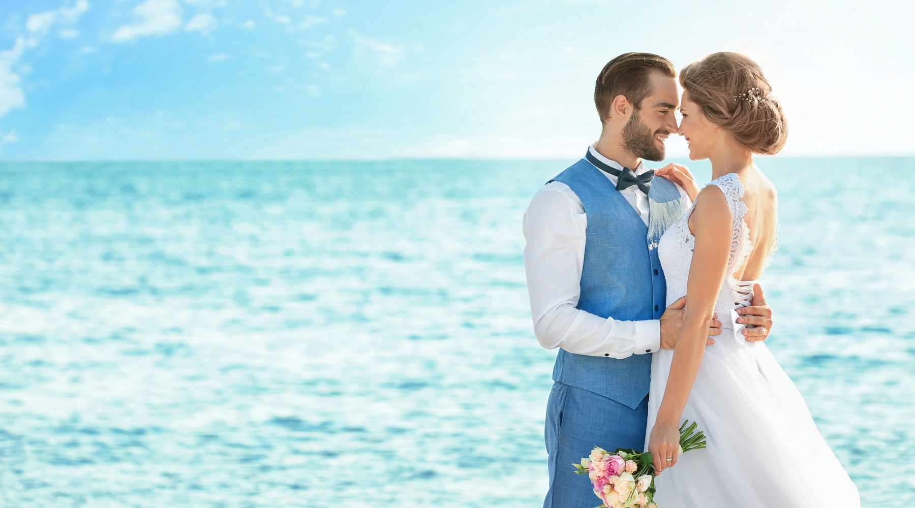 A close-up of a man wearing a blue suit with a vest holding a woman wearing a white wedding dress by the ocean.