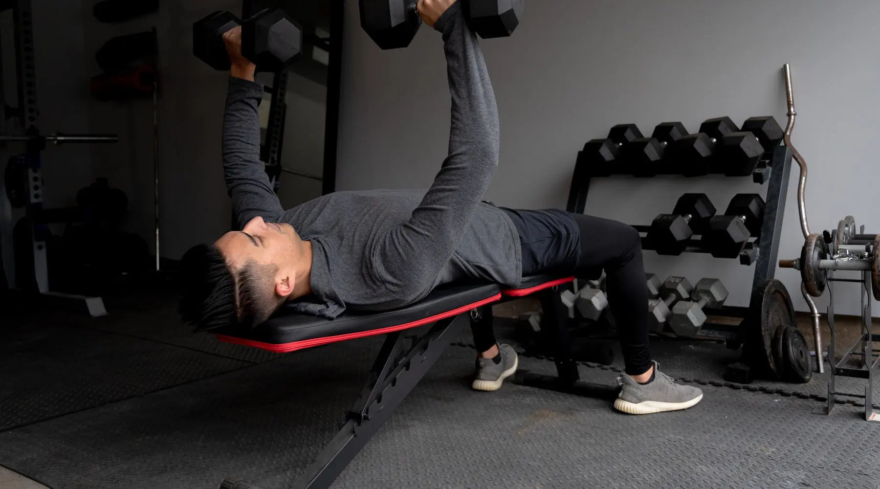 A young adult lying on his back on top of a black bench placed over black foam floor while holding two dumbbells.