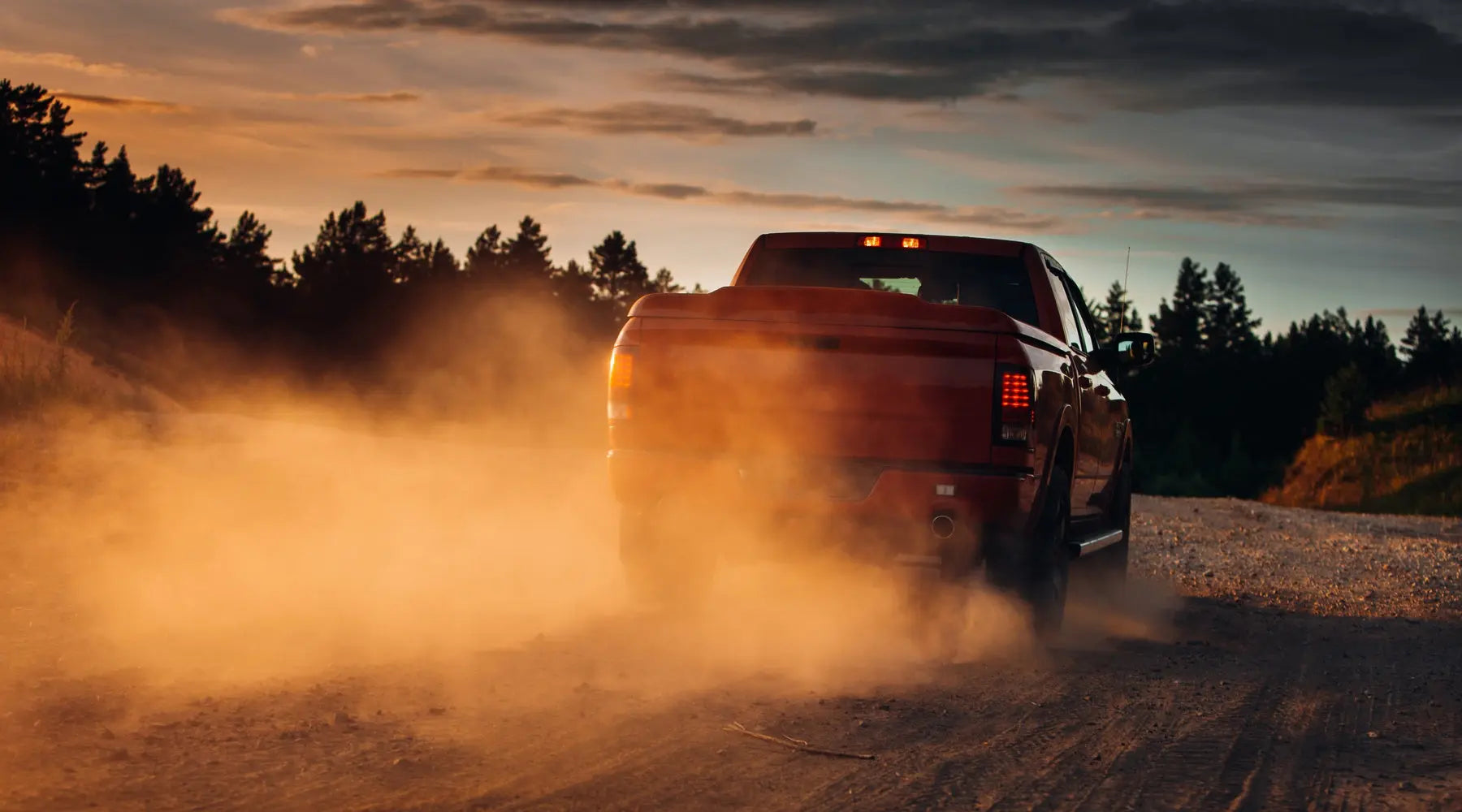 A red pickup truck with a large bed driving through a dirt road, leaving a dust cloud behind during a sunset.