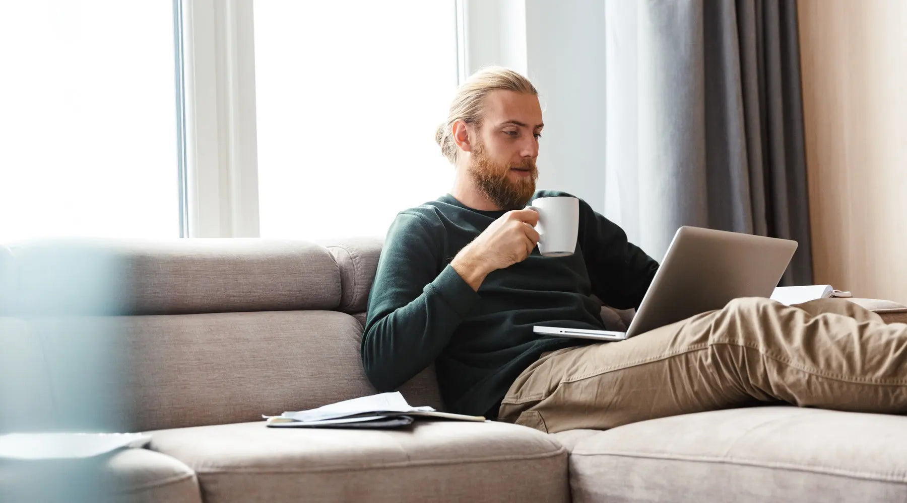 A bearded man sits on the couch with his feet up. His laptop is in his lap and he's holding a white coffee mug.