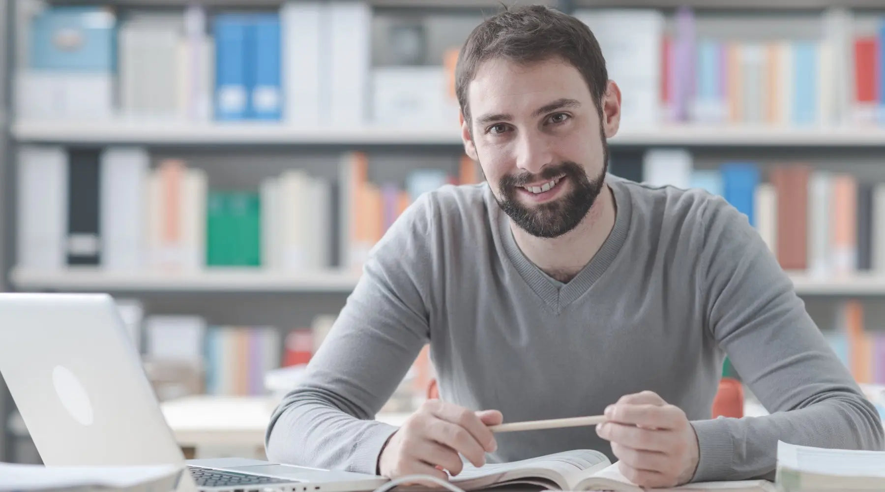 A young man smiles while surrounded by a laptop, books, and other office essentials. A large bookshelf is behind him.