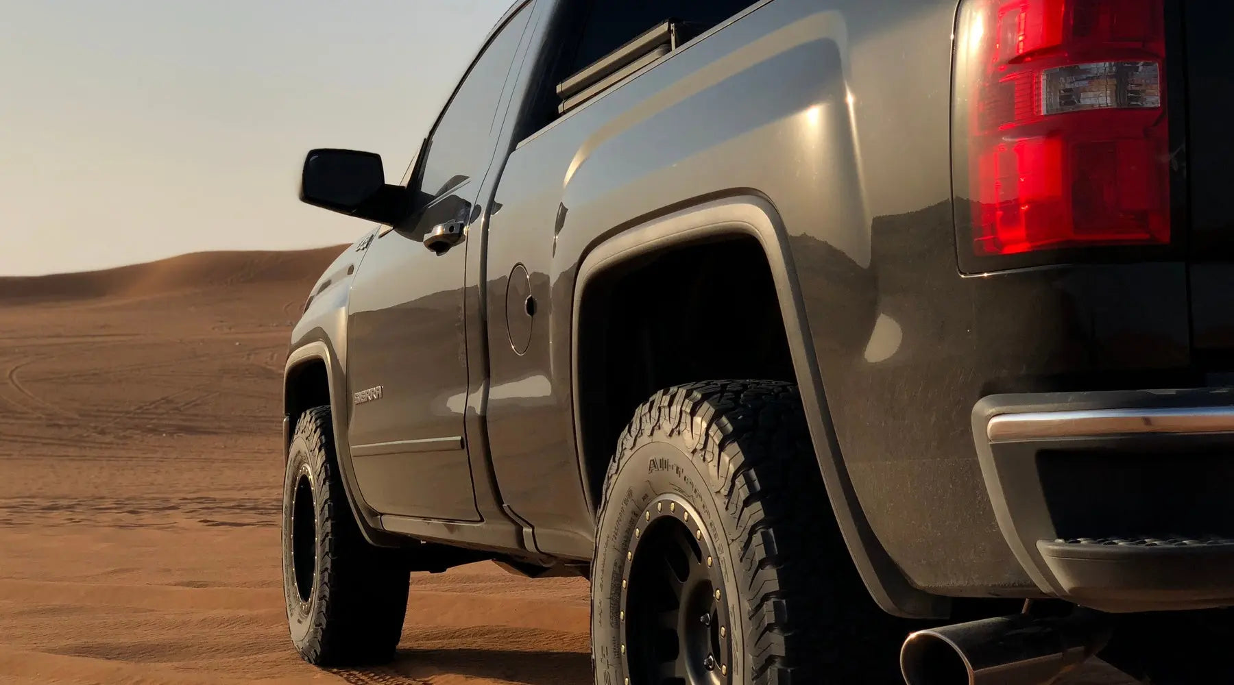 A close-up shows a black pickup truck on a sandy surface, with hills in the background. Tire tracks appear in the sand.