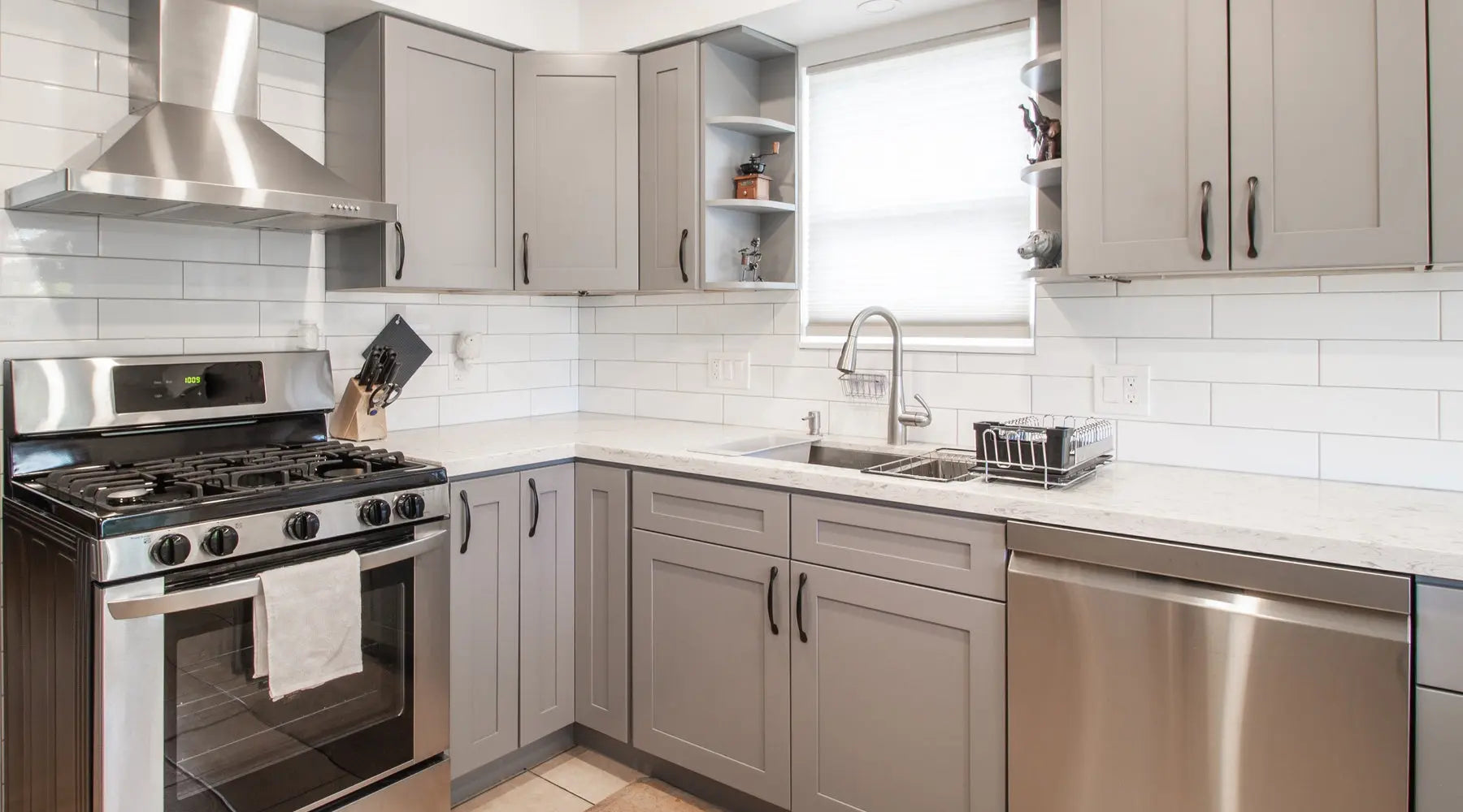 A small, modern kitchen with gray cabinets and black hardware. There are some open shelves and stainless steel appliances.