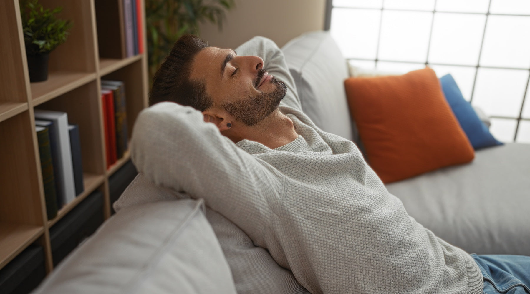 A man with short hair is resting on a couch in a living room. There is a window behind him with light coming in.