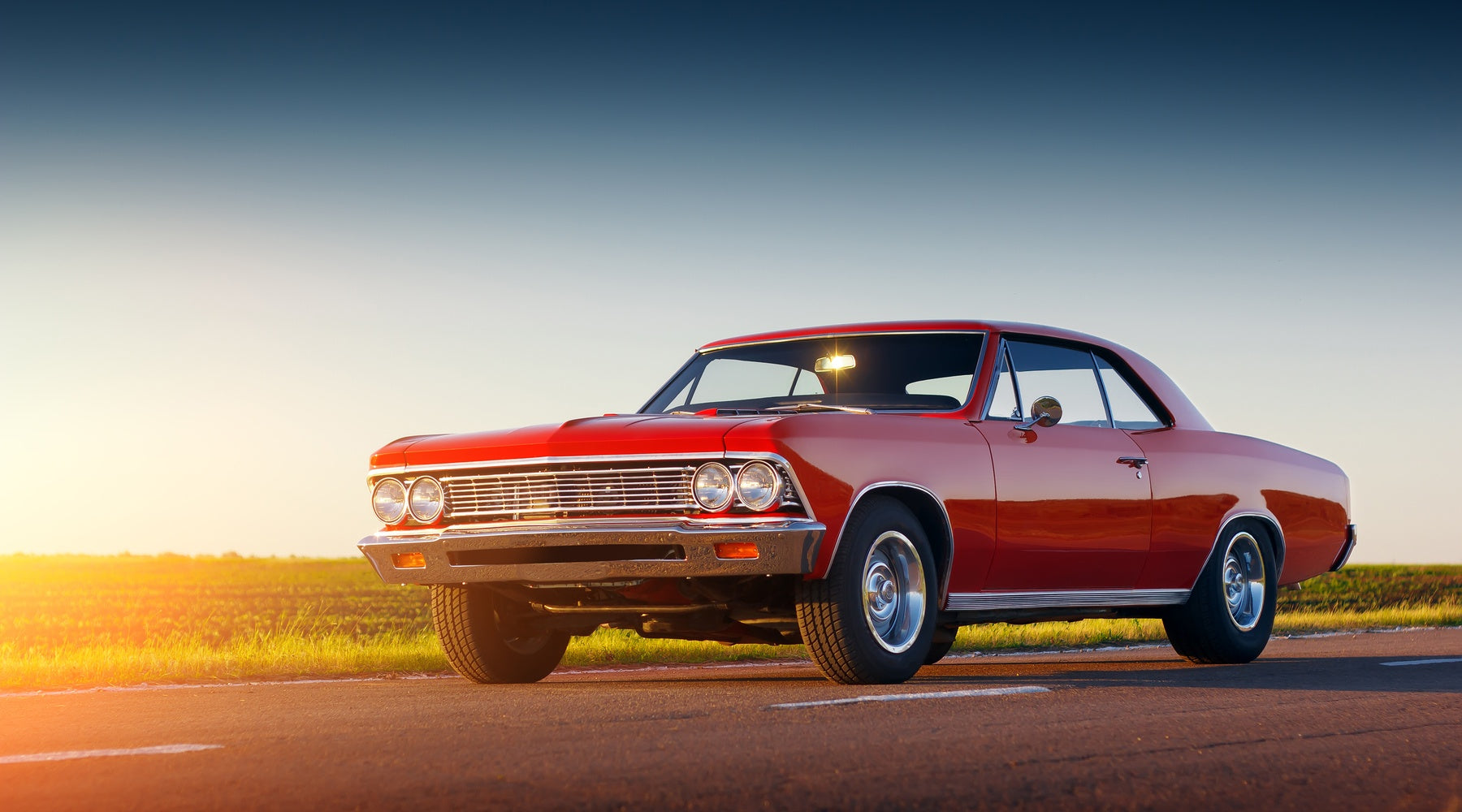 Red classic car parked on open road at sunset, with warm light reflecting on body and clear sky over rural landscape.