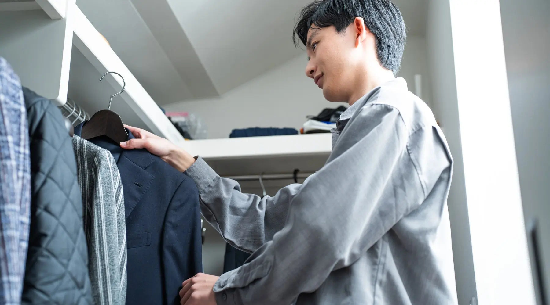 A young man in a gray long-sleeve shirt hanging a suit jacket on a wooden hanger on the clothing rod inside a closet.