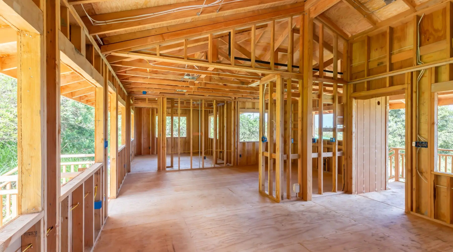 The interior of a large room under construction with large windows, exposed wooden beams, and electrical wires.