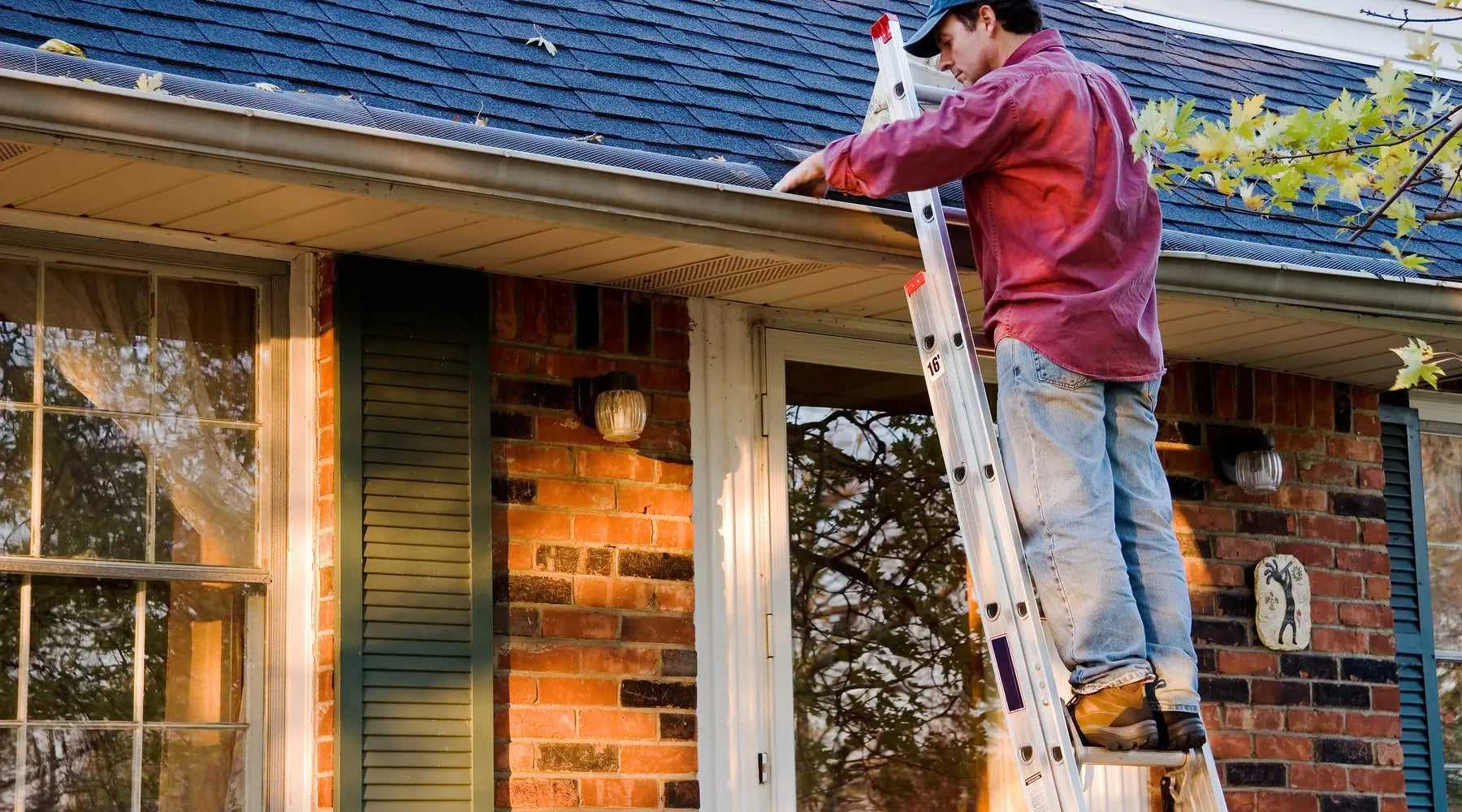A man wearing a red shirt and a blue cap stands on a ladder near tree branches, reaching into the gutters of a home.