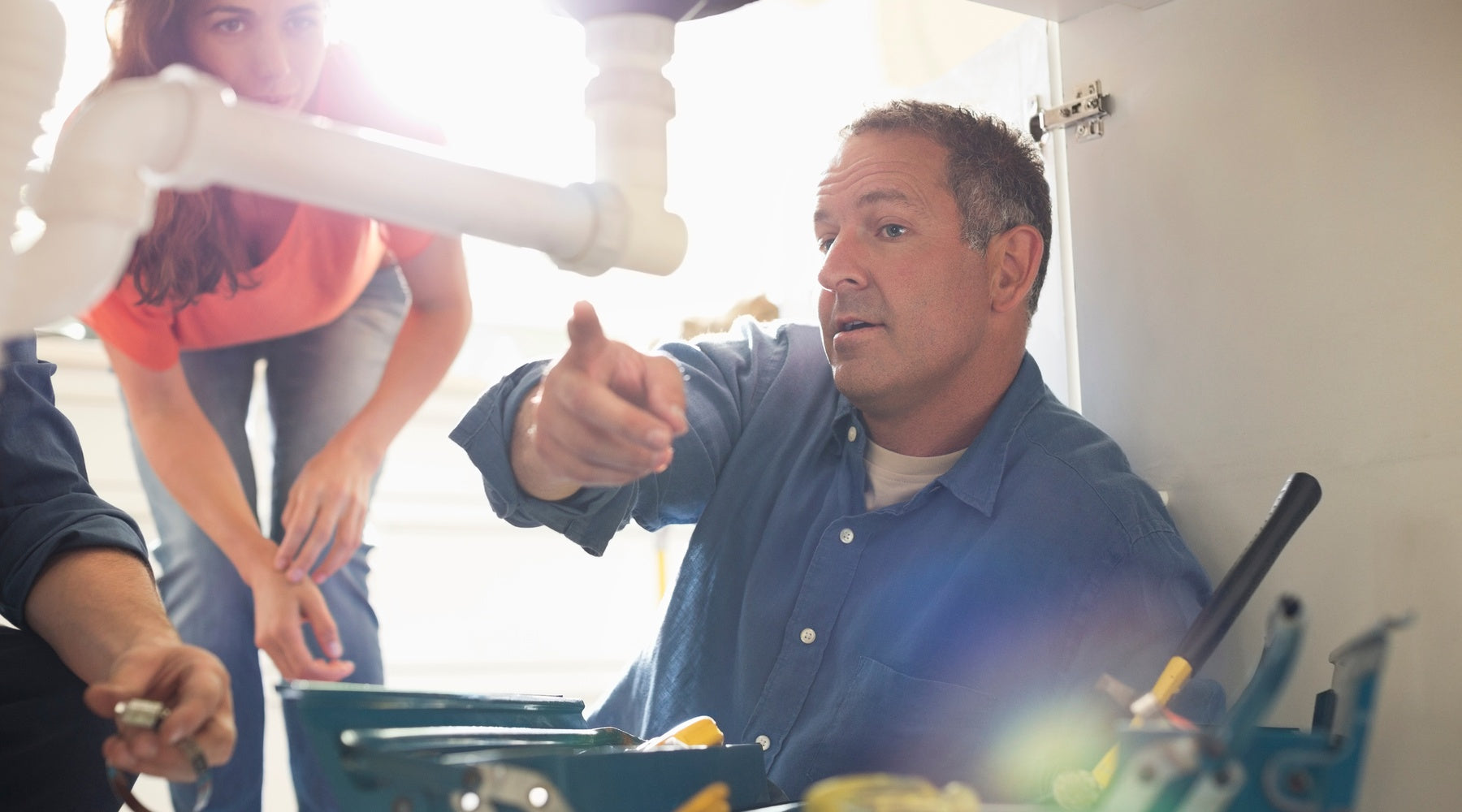 A plumber pointing out an issue with some pipes that are underneath a sink to a homeowner who is standing next to him.