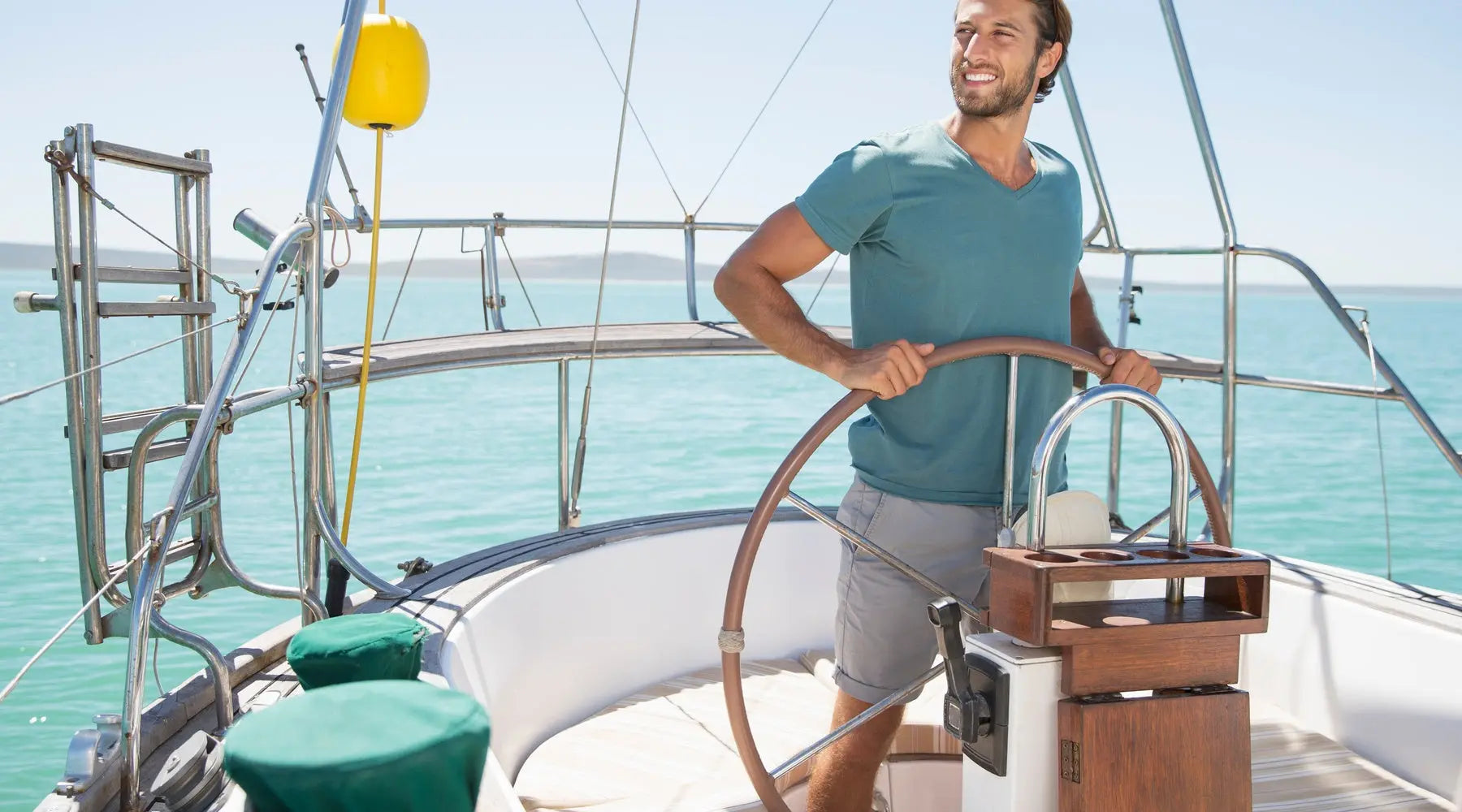 A single man steers a boat's wheel as he smiles and looks off into the distance. It's sunny outside.