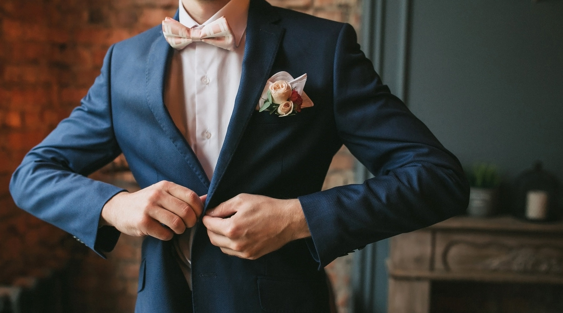 A Caucasian man buttons his navy coat and wears a pink bow tie in front of a brick wall and fireplace.