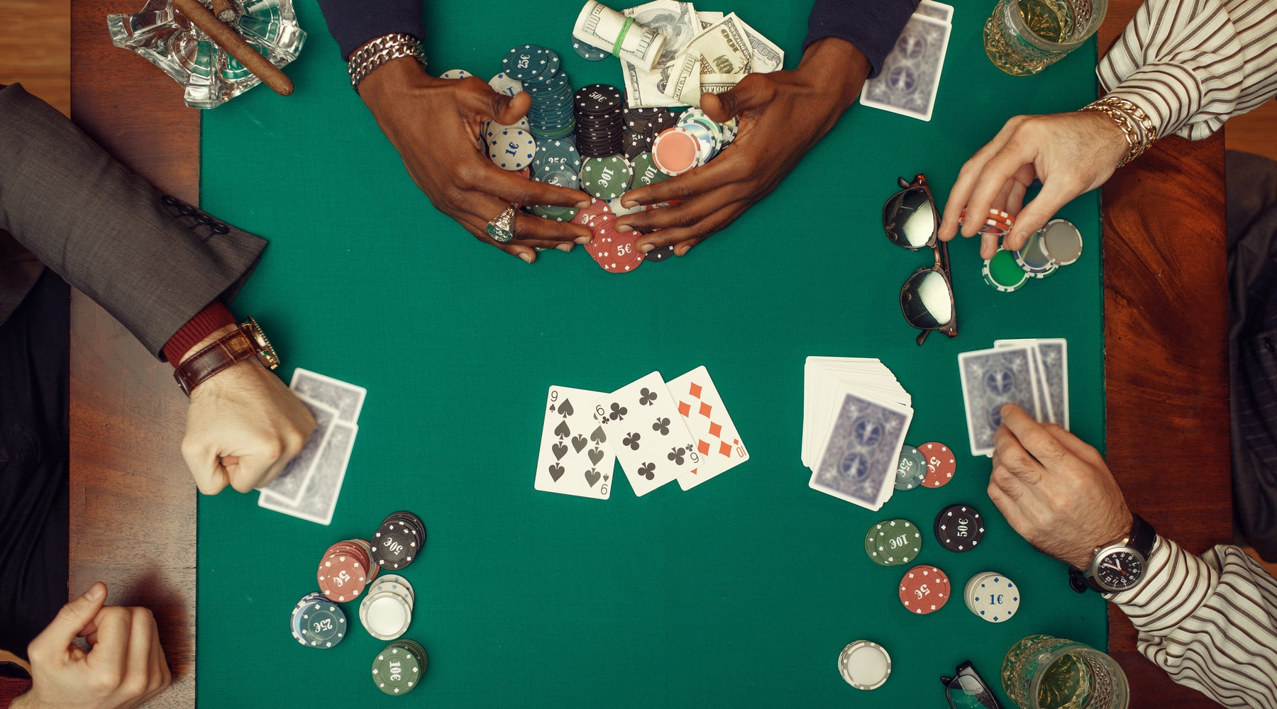 Four people playing poker at a green table with cards, chips, cash, and drinks arranged during a competitive game.