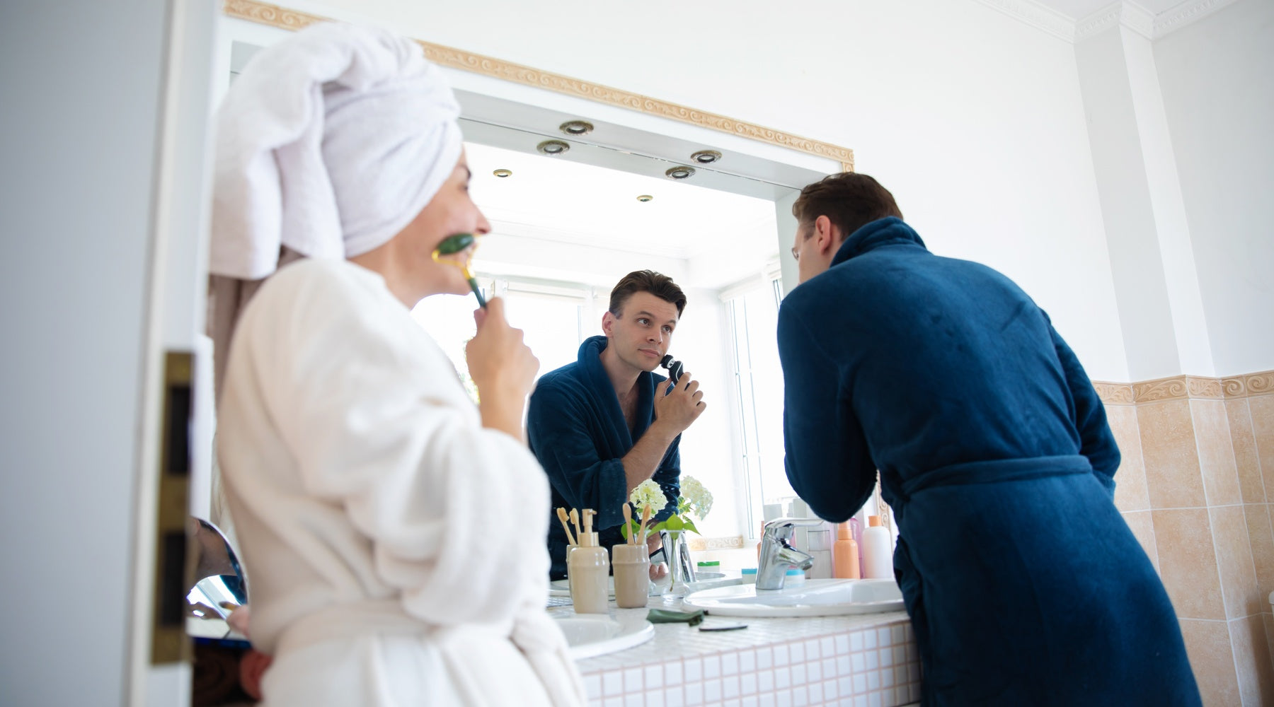 Two people wearing robes in their bathroom at home. Both people shave their faces in front of a large mirror.