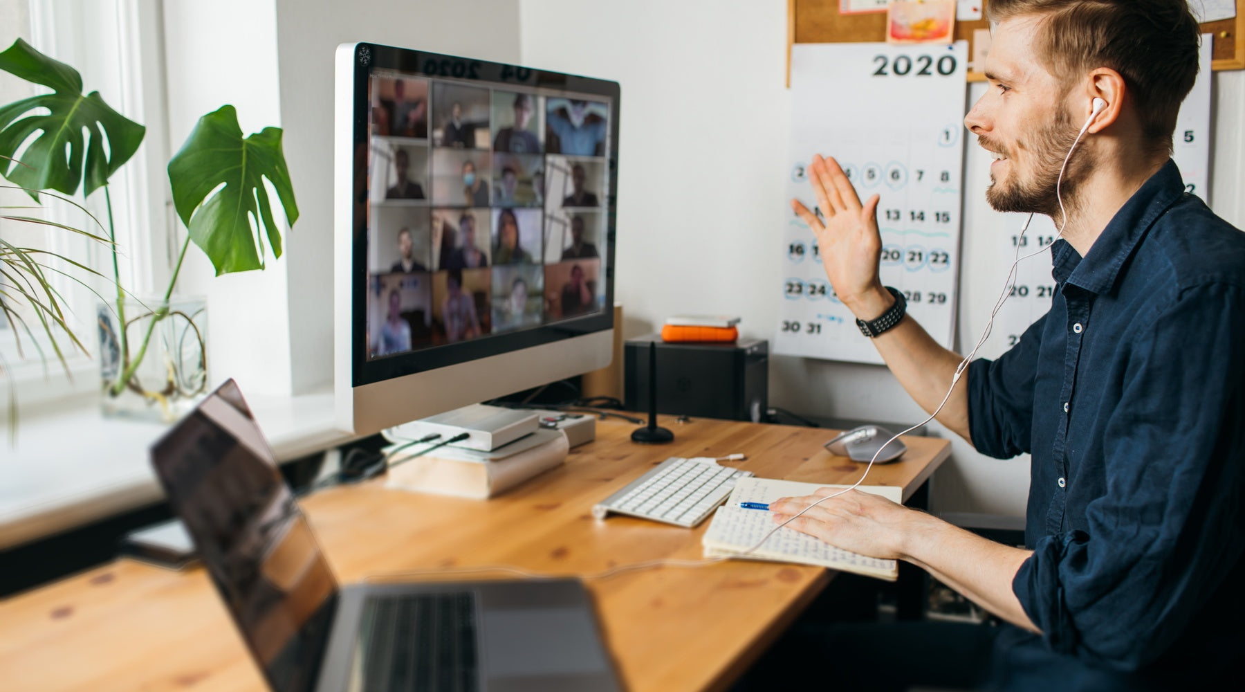 A remote work creative professional sits at his desk with a laptop and an external monitor. He waves to the team meeting.
