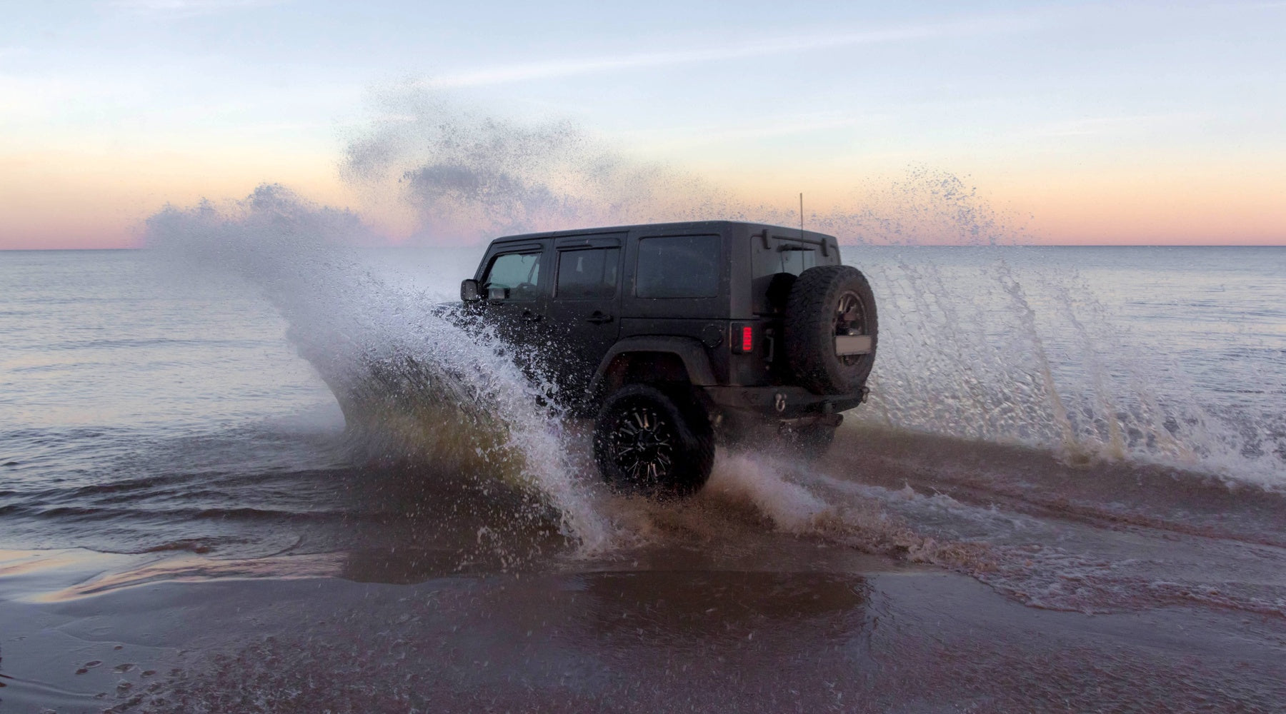 A black Jeep on the beach splashing water as it drives through the shoreline. The sun is setting in the background.