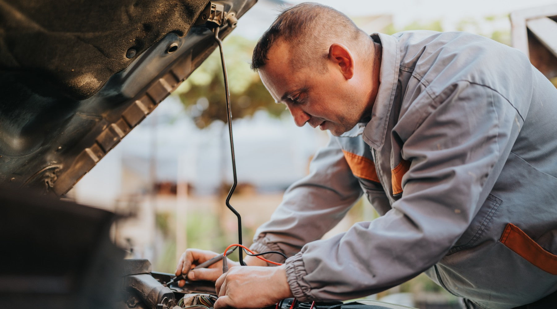 A car owner wearing a jacket is using a small tool to make electrical repairs under the hood of their vehicle.