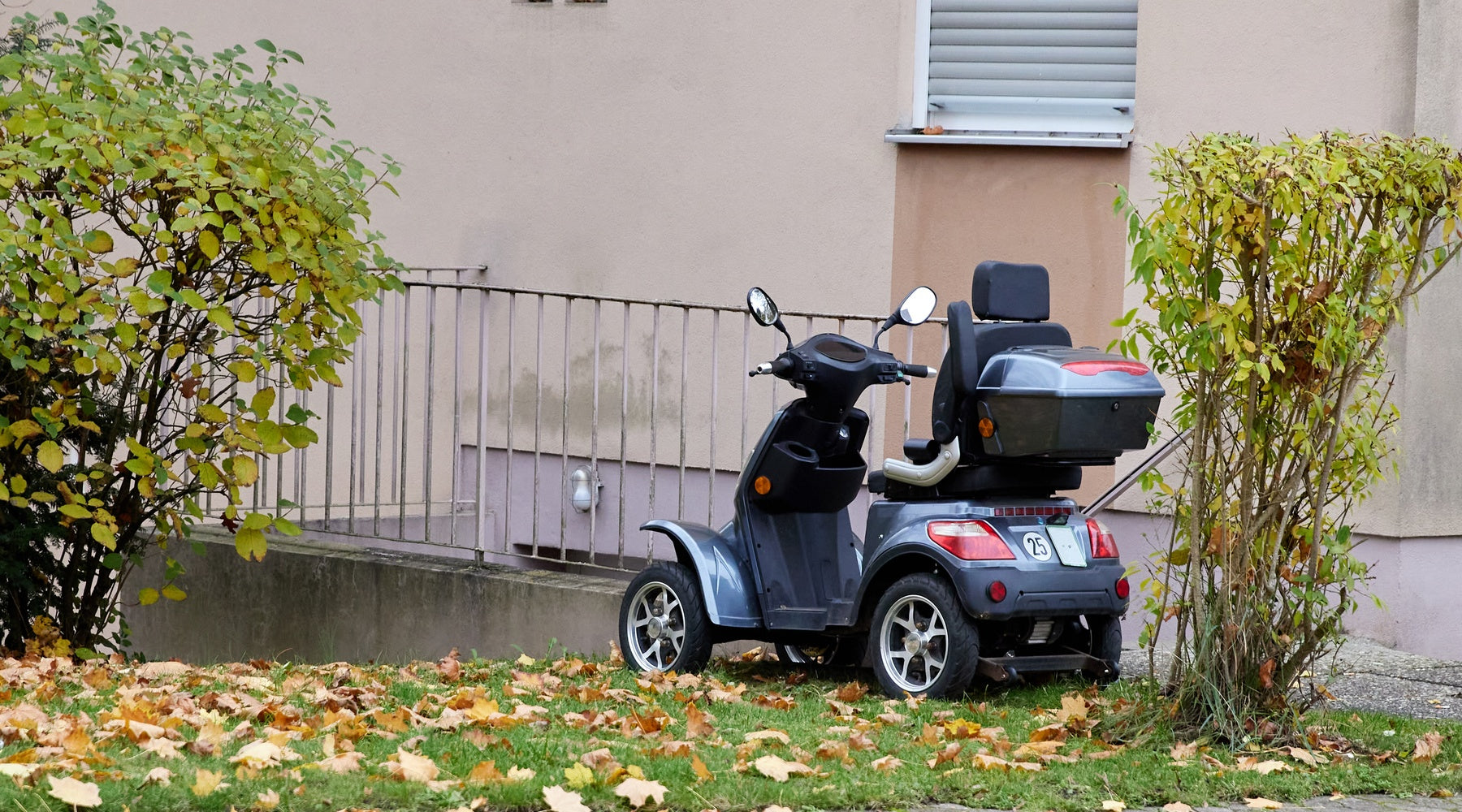 An empty mobility scooter parked outside near a metal railing. Two bushes with green leaves are nearby.