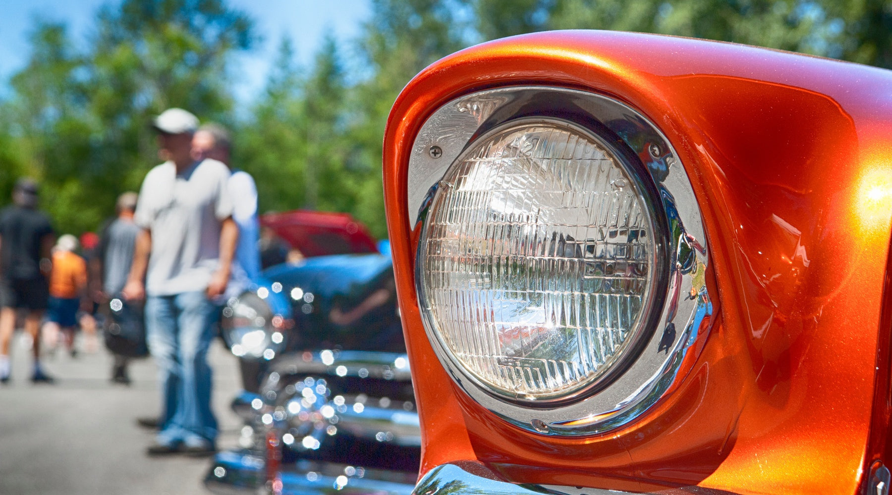An orange classic car fills the frame while visitors browse polished vintage cars at a sunny outdoor meet.