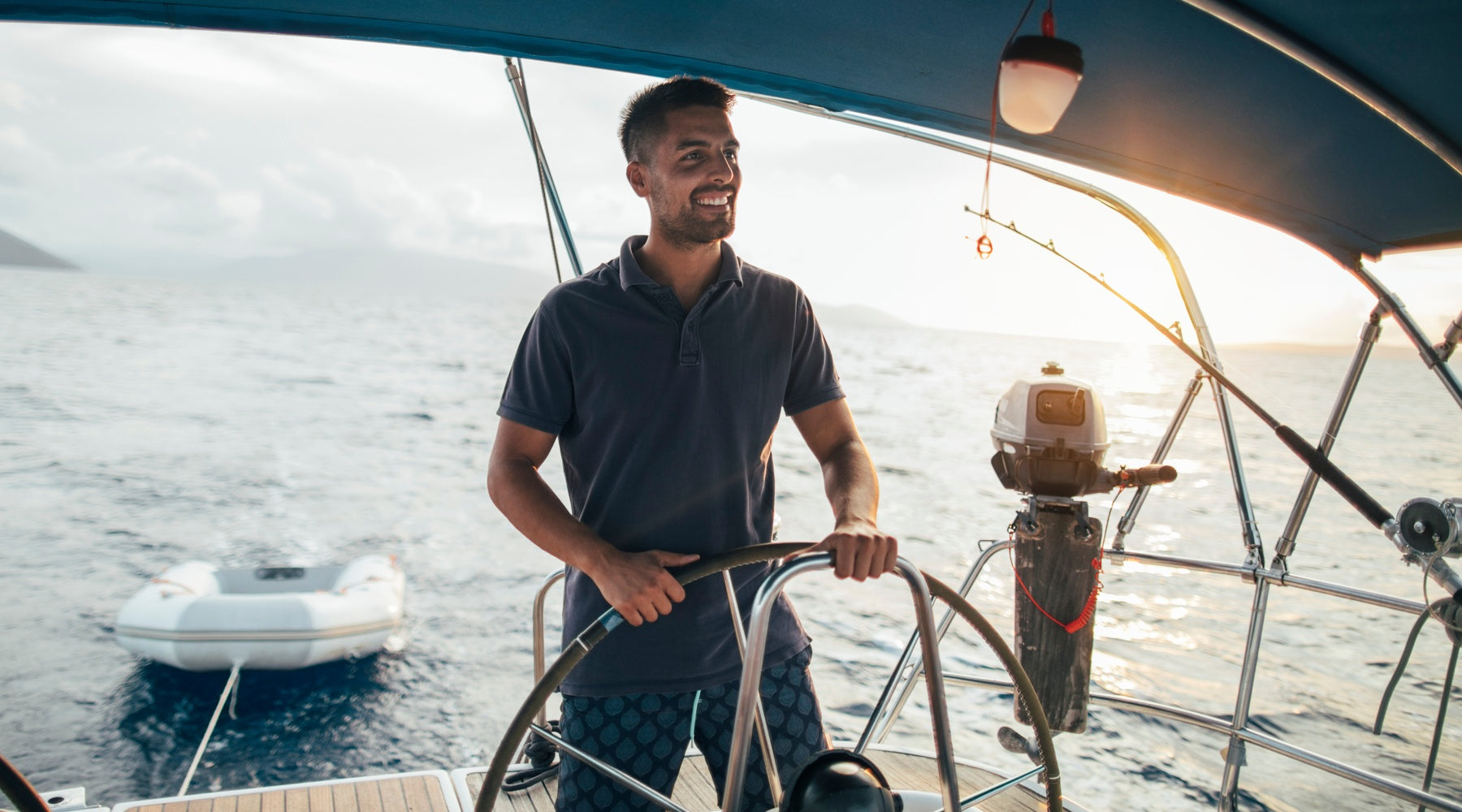 A young man wearing a polo shirt smiles while holding the helm of a sailboat. The open water is in the background.