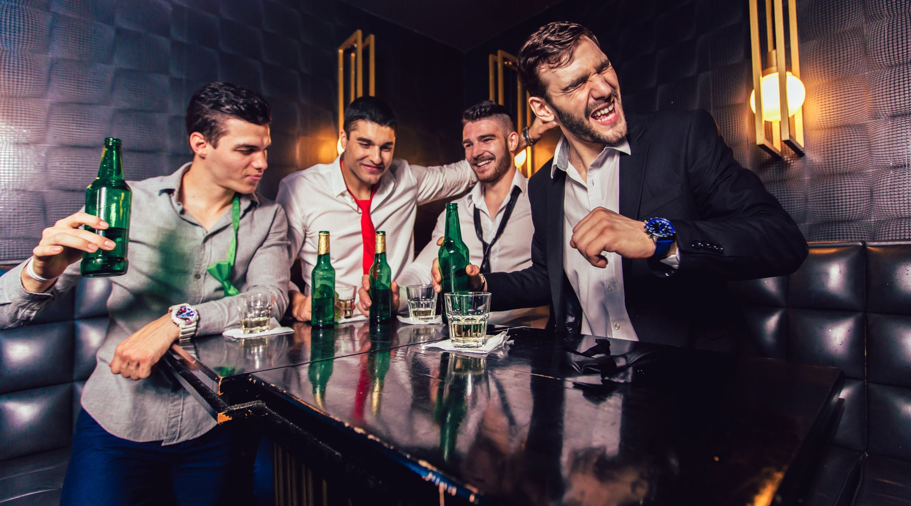 Four men party together in a dark lounge booth with beer bottles and glasses as one man laughs across the table.