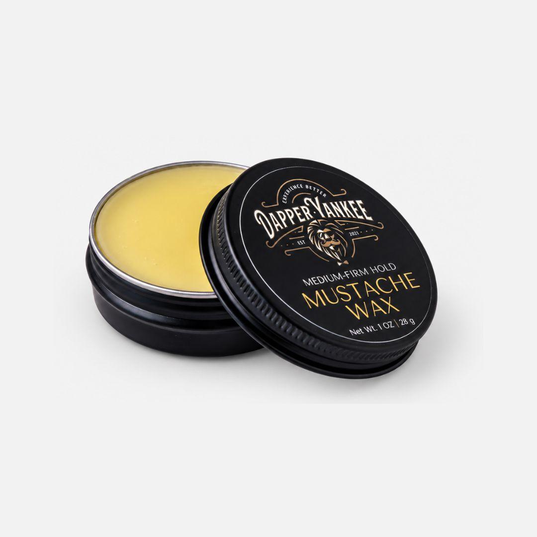An open round black tin of Dapper Yankee Mustache Wax with pale yellow wax visible, next to its black lid featuring gold lettering, and the Dapper Yankee logo.