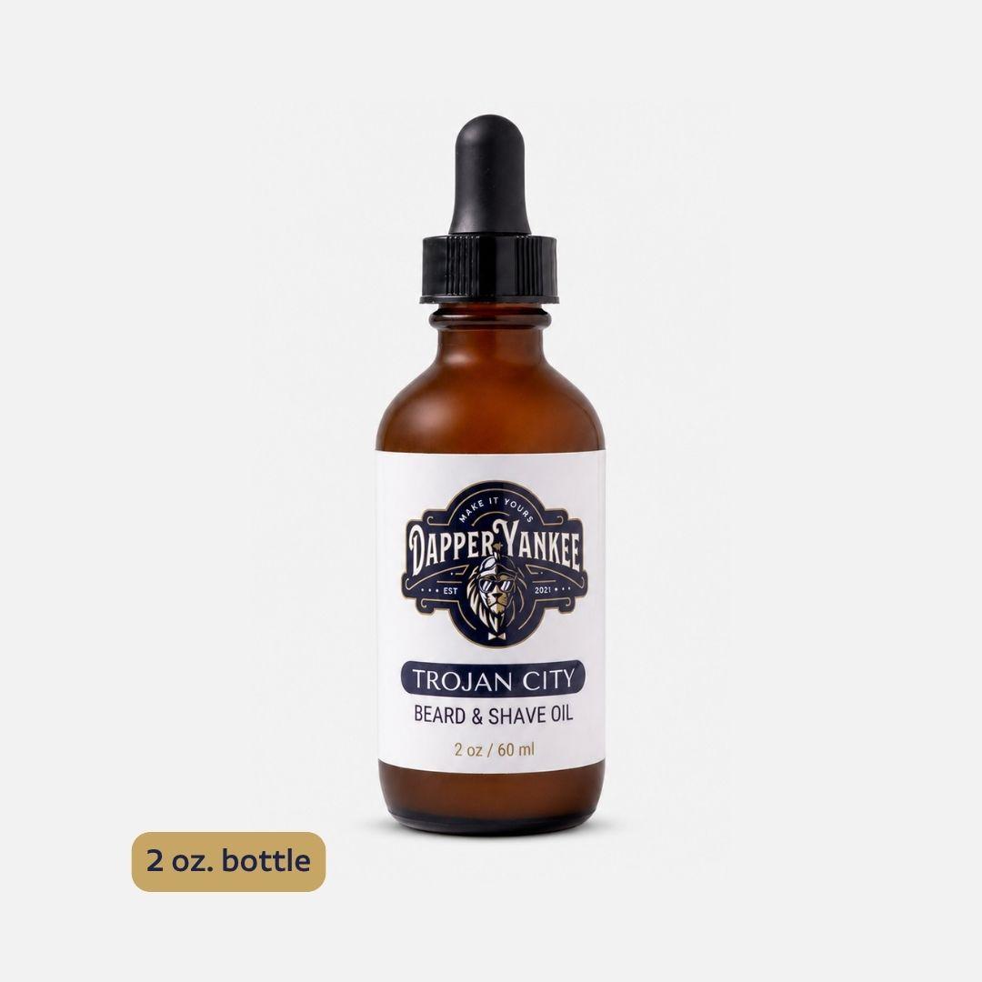 A 2 oz amber glass dropper bottle of Dapper Yankee Trojan City Beard & Shave Oil on a light gray background, with a gold '2 oz. bottle' label overlay in the bottom left.