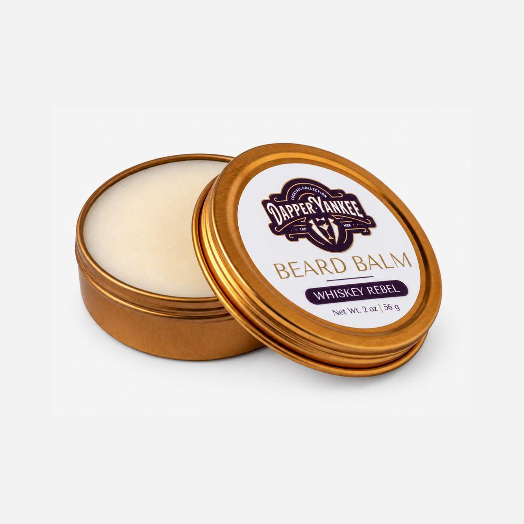 An open round metal tin of Dapper Yankee Beard Balm with the lid leaning against it, showing the cream-colored balm inside. The lid features a white label with the Dapper Yankee logo and gold and purple text reading 'Beard Balm Whiskey Rebel'.