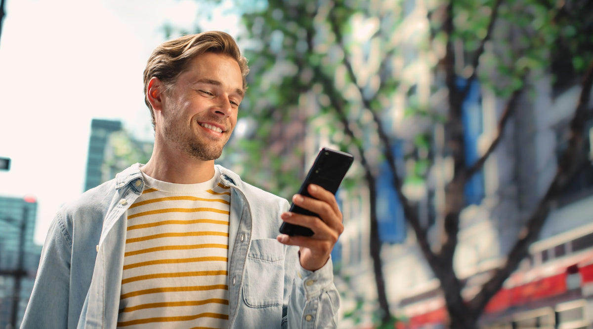 A smiling man in a striped shirt checks a smartphone while standing on a sunny, tree-lined city street.