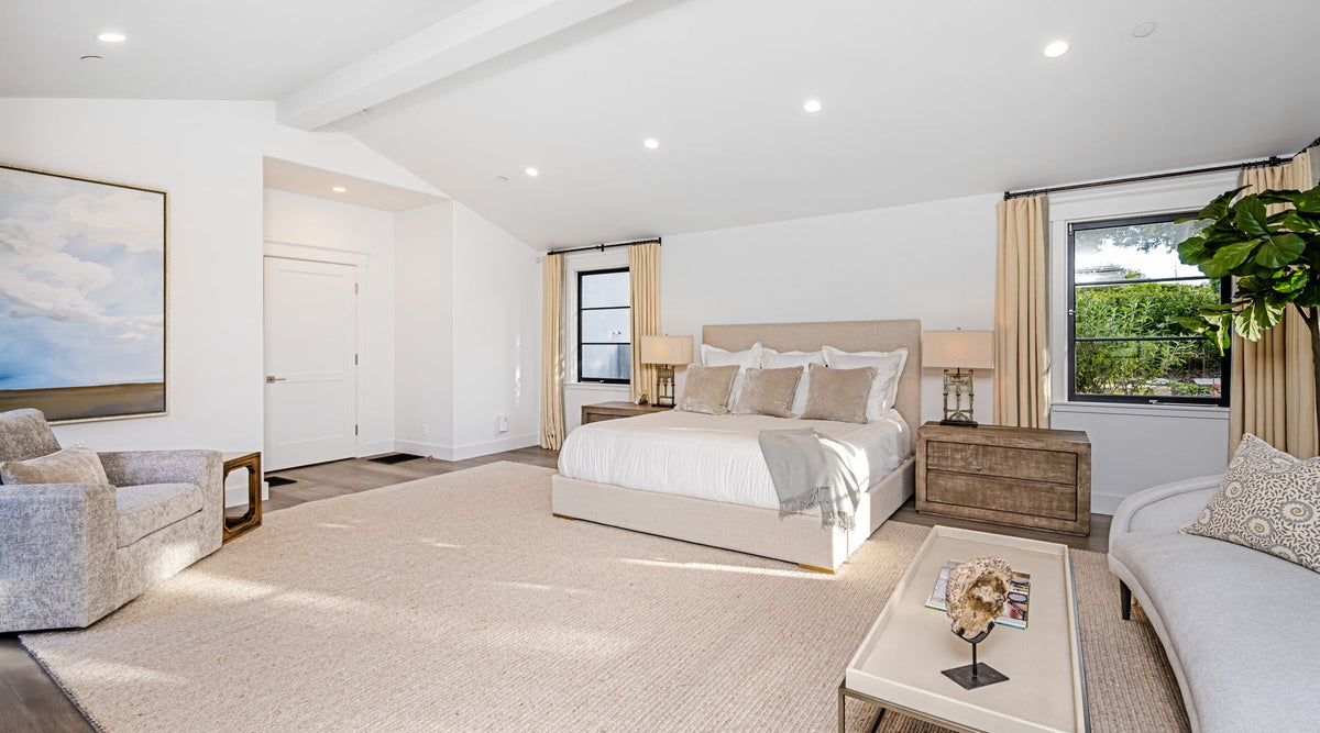 A large bedroom with a bed, a beige rug on hardwood floors, a gray armchair, and a small sofa by a coffee table.