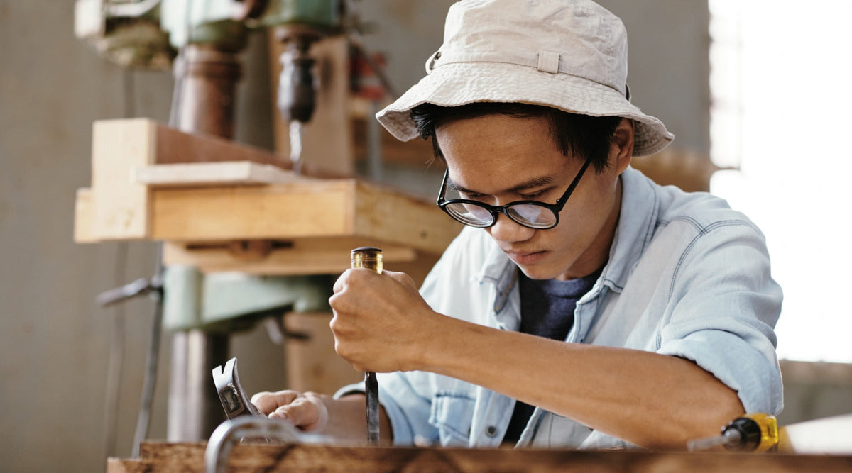 A man wearing glasses and a bucket hat using a chisel and hammer on a wooden piece inside a workshop with tools nearby.