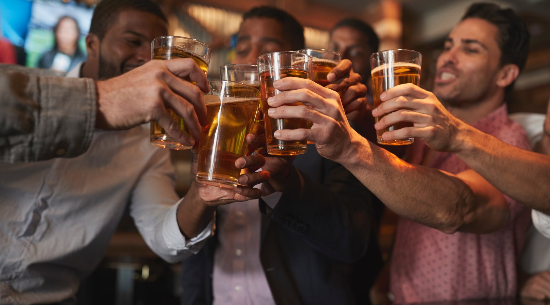 A group of diverse young male friends toasting full pint glasses of beer in a bar with TVs in the background.