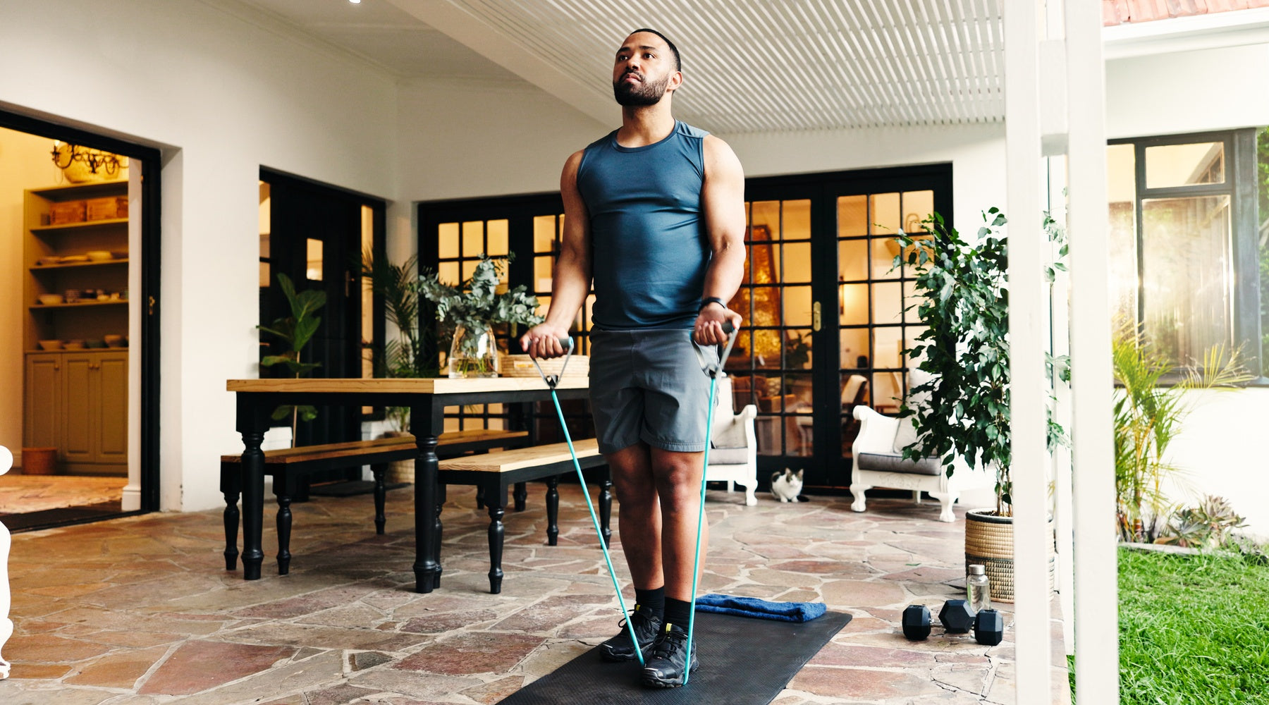 A man standing on a yoga mat, holding an elastic workout band with two hands under his foot in an outdoor space.
