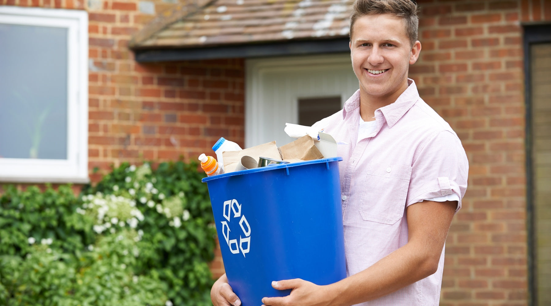 A young man smiles as he carries a recycling trash container away from his house. He wears a button-down shirt.