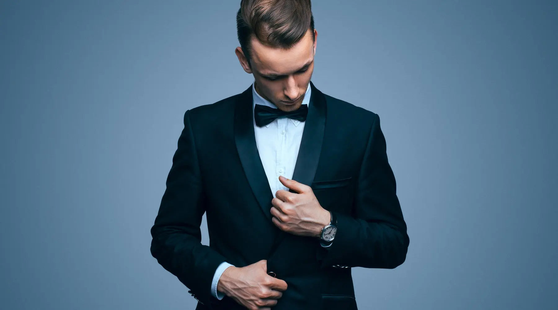A young man adjusts the lapels on his tuxedo. He's wearing a black bow tie and a silver watch.