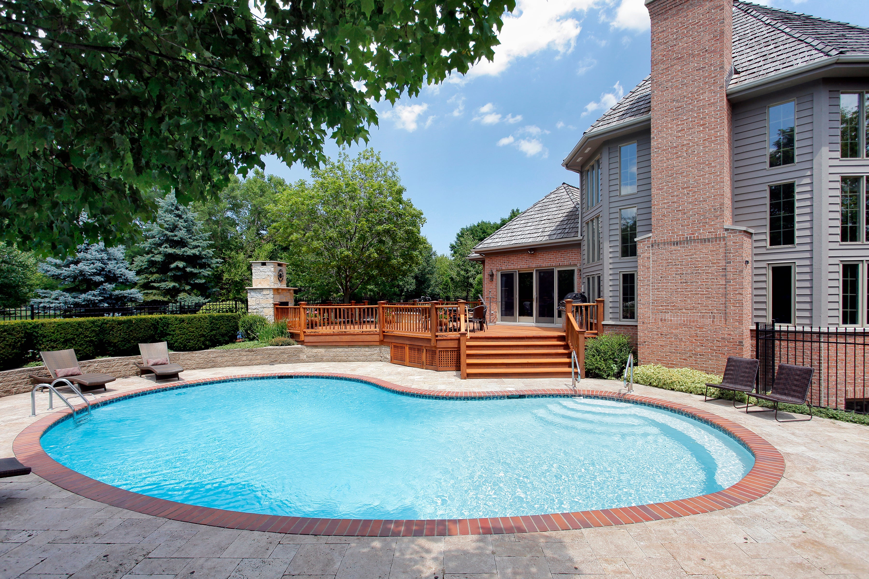 A backyard with a concrete paved surface around a curved pool. A wooden deck is attached to the backside of the house.