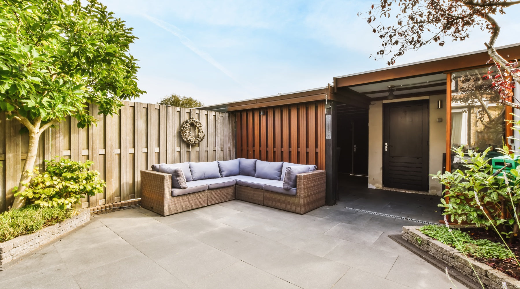A large backyard with a wooden fence surrounding it, a sectional couch, a small pergola, concrete floors, and greenery.