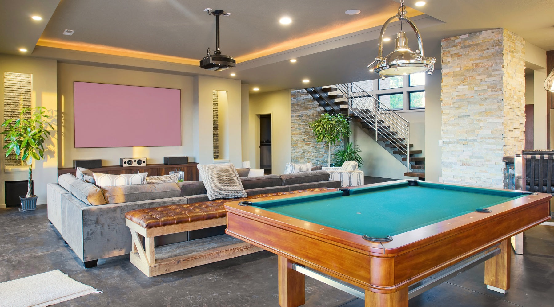 A luxury basement man cave features a wooden pool table, a sectional sofa, and a projector screen on the wall.