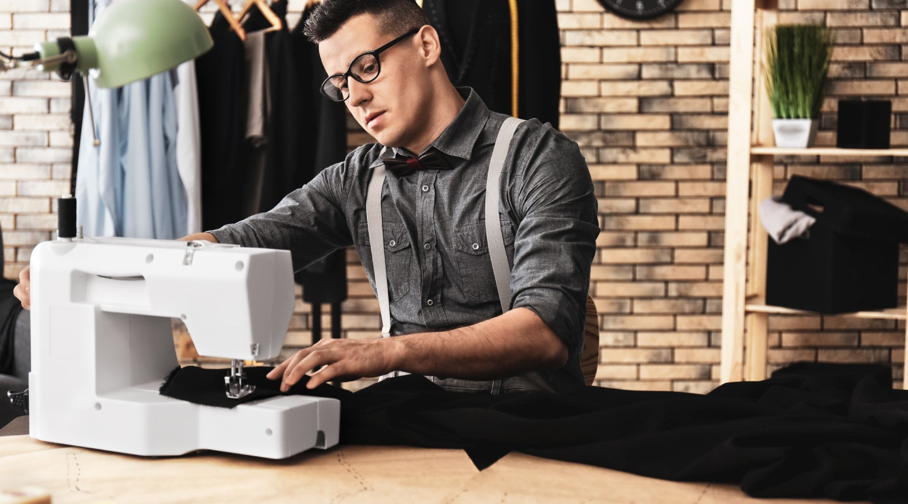 A man running black fabric through a white sewing machine. He sits at a table in front of a brick wall and rack of clothes.