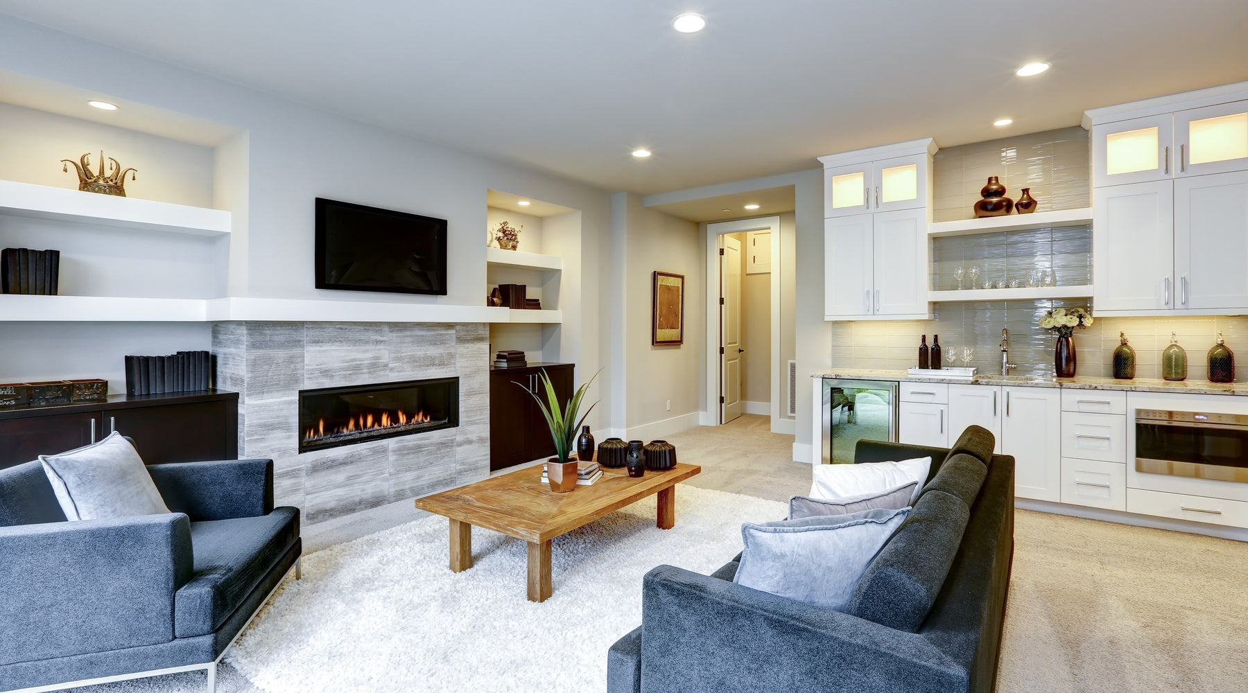 A living area has a fireplace, a TV, and two gray sofas. White cabinets, a sink, and a drink fridge line the wall.