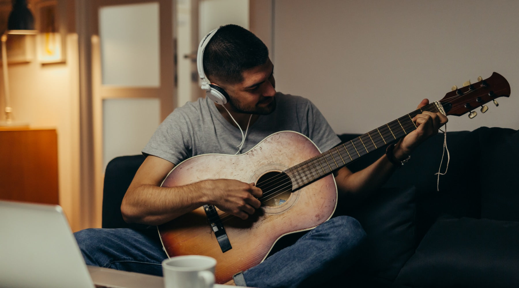 A man playing a guitar while sitting on a couch in front of a laptop. He is wearing white headphones on his head.