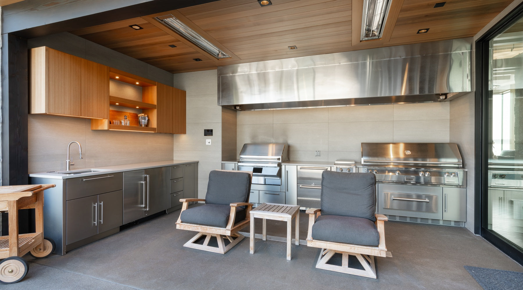 A luxury outdoor kitchen features a large built-in grill, a beverage cooler, a sink, two lounge chairs, and wooden cabinets.