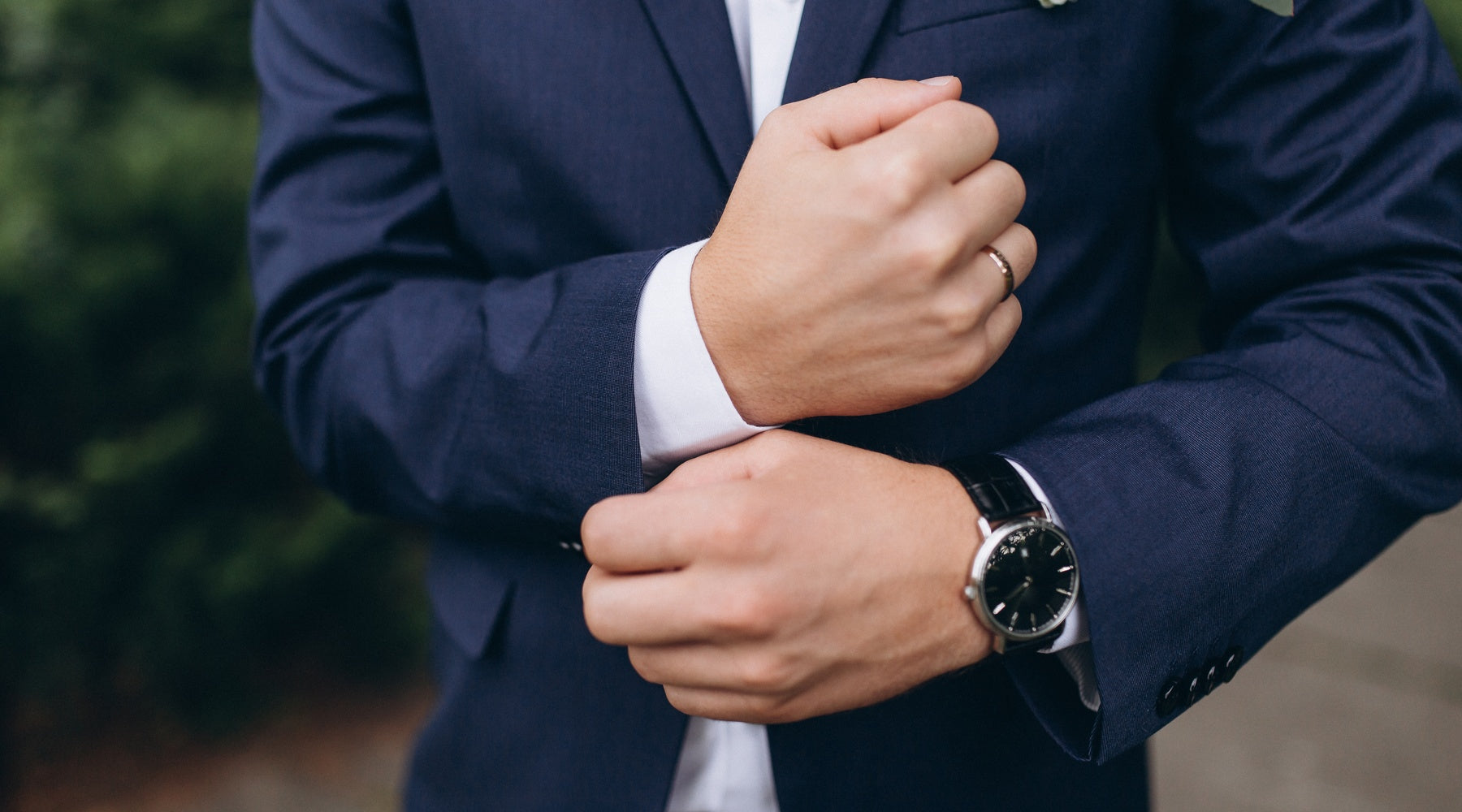 A man is adjusting his cuffs. He’s wearing a suit with a tie and he has a watch strapped to this left wrist.